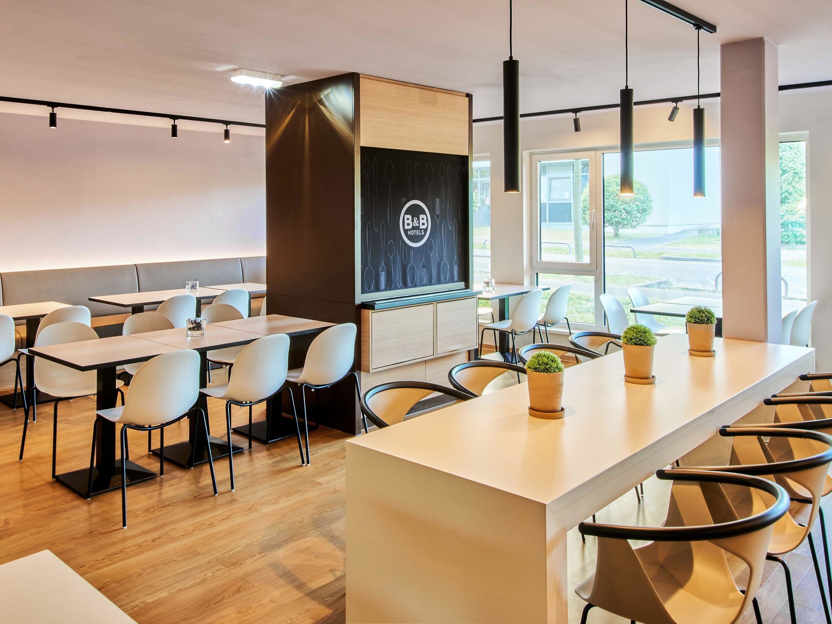 Eine moderne Hotelhalle mit einer langen Rezeption, mehreren Tischen und Stühlen und großen Fenstern mit Blick ins Freie.