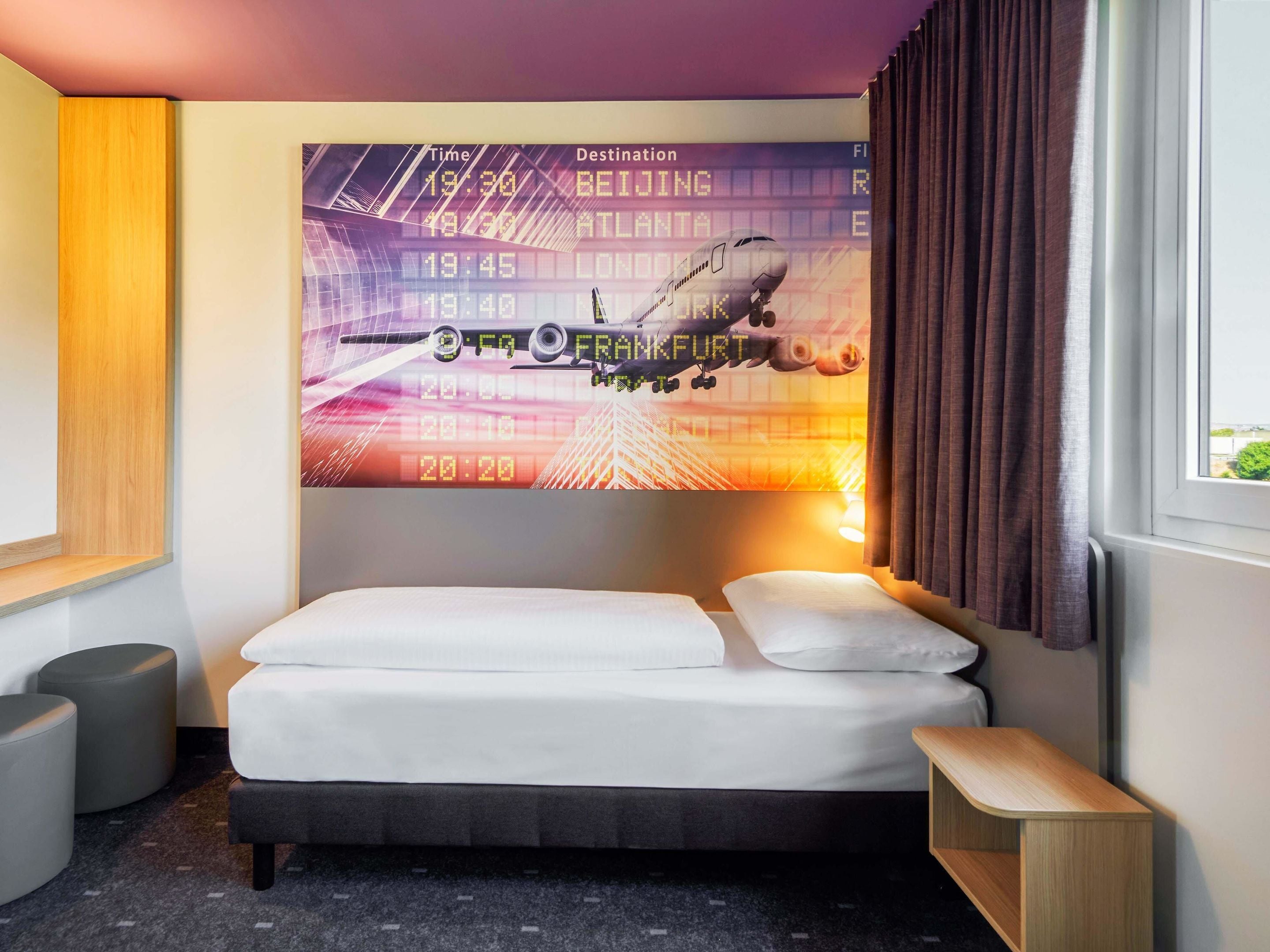 A hotel room with a bed, a small bedside table, a wall-mounted TV displaying flight information, and a window with curtains.