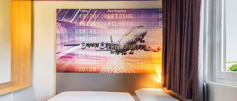 Habitación de hotel con cama, una mesita de noche, televisor empotrado mostrando información de vuelos y una ventana con cortinas.