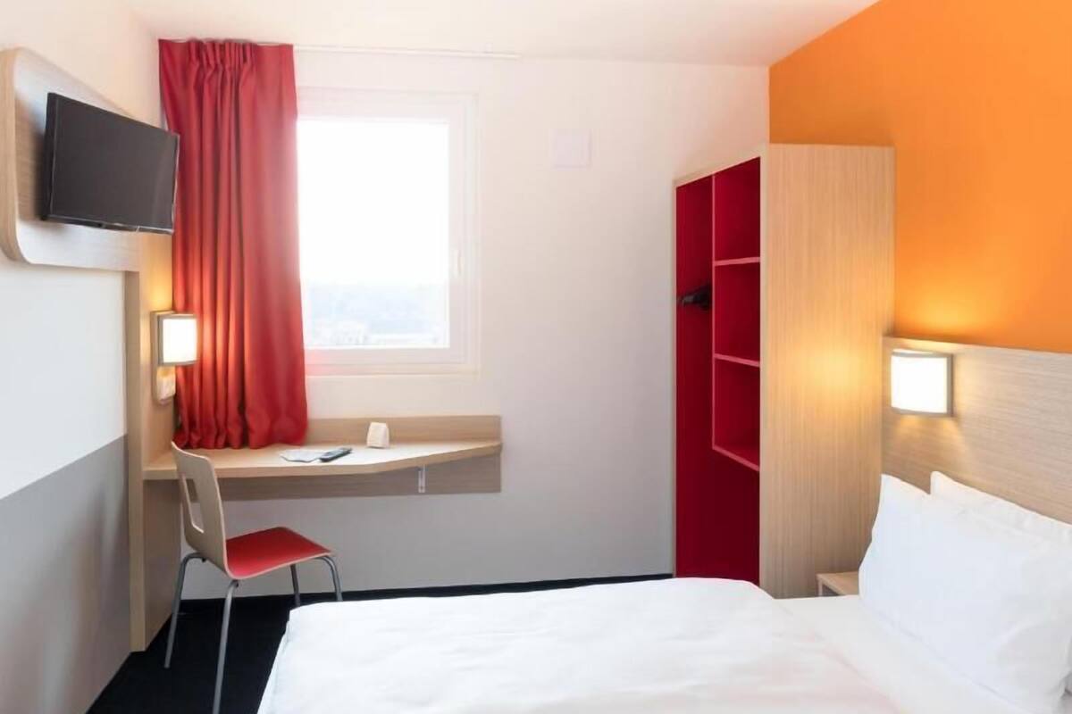 A hotel room with a bed, a desk with a chair, a television, and a window with curtains.