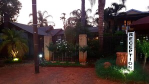 Zona de recepción con un cartel, paseo empedrado y palmeras.