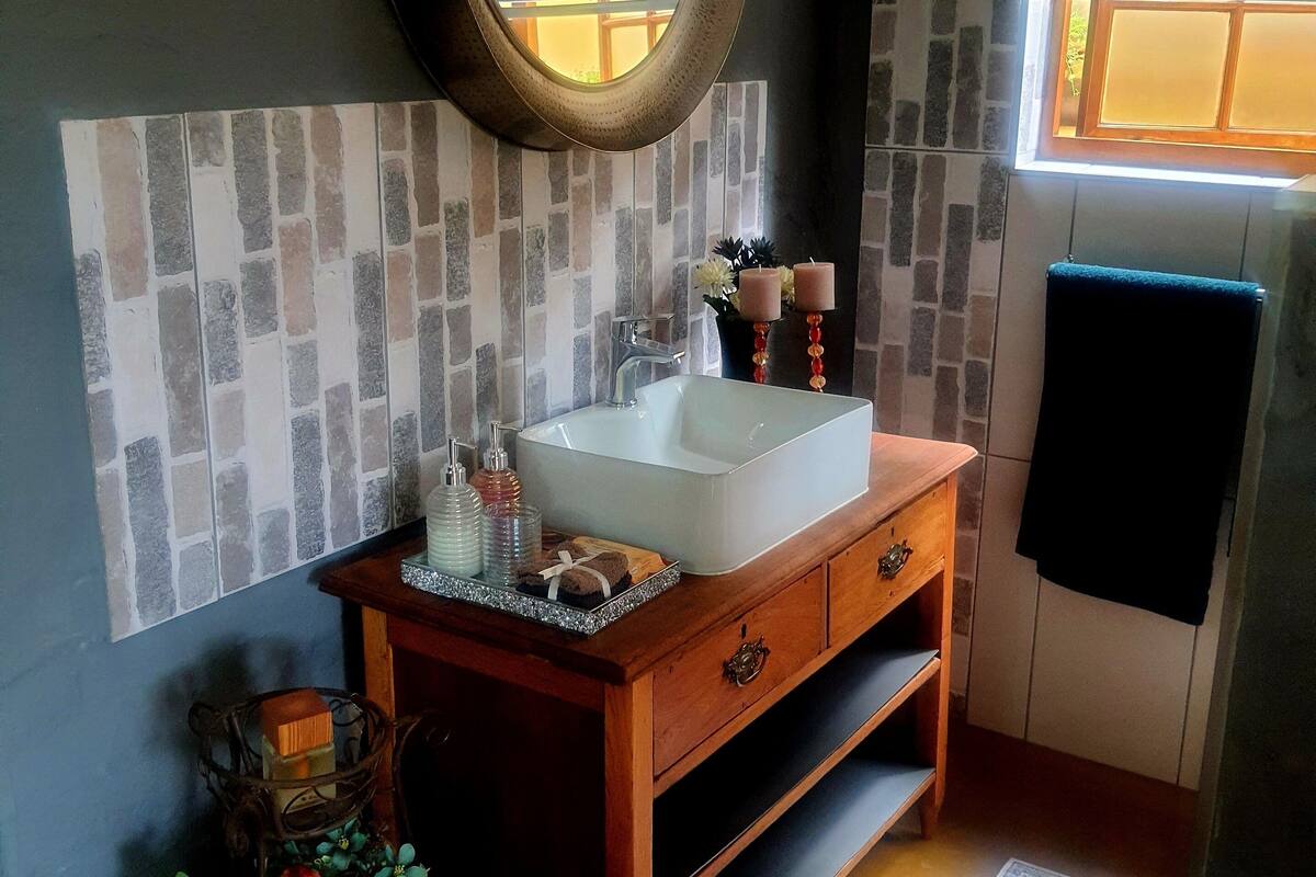 Une salle de bain avec un lavabo blanc, un meuble-lavabo en bois et un miroir. On y trouve une fenêtre avec des stores, une serviette accrochée au mur et une plante décorative.
