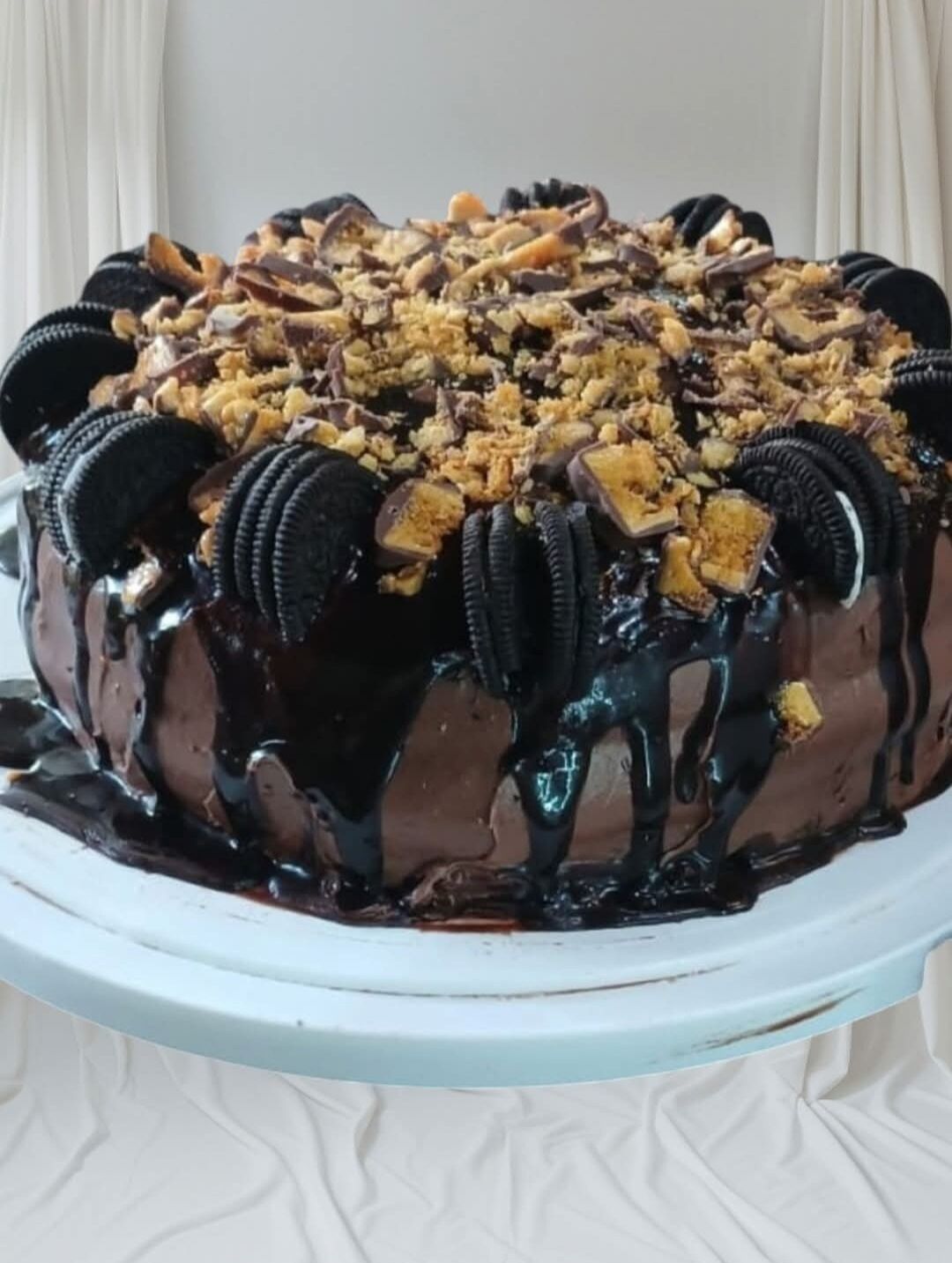 A chocolate cake with Oreo cookies and crushed nuts on a white cake stand.