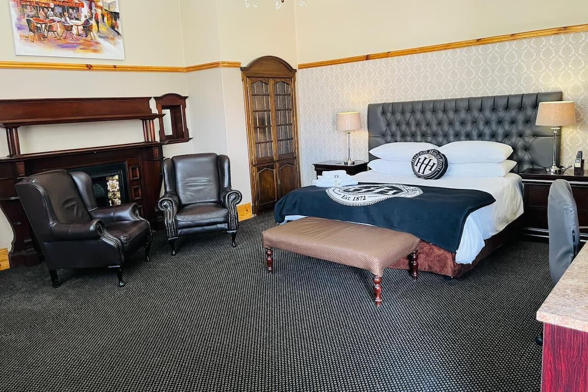 A hotel room with a large bed, two armchairs, a bench, a fireplace, and a chandelier.