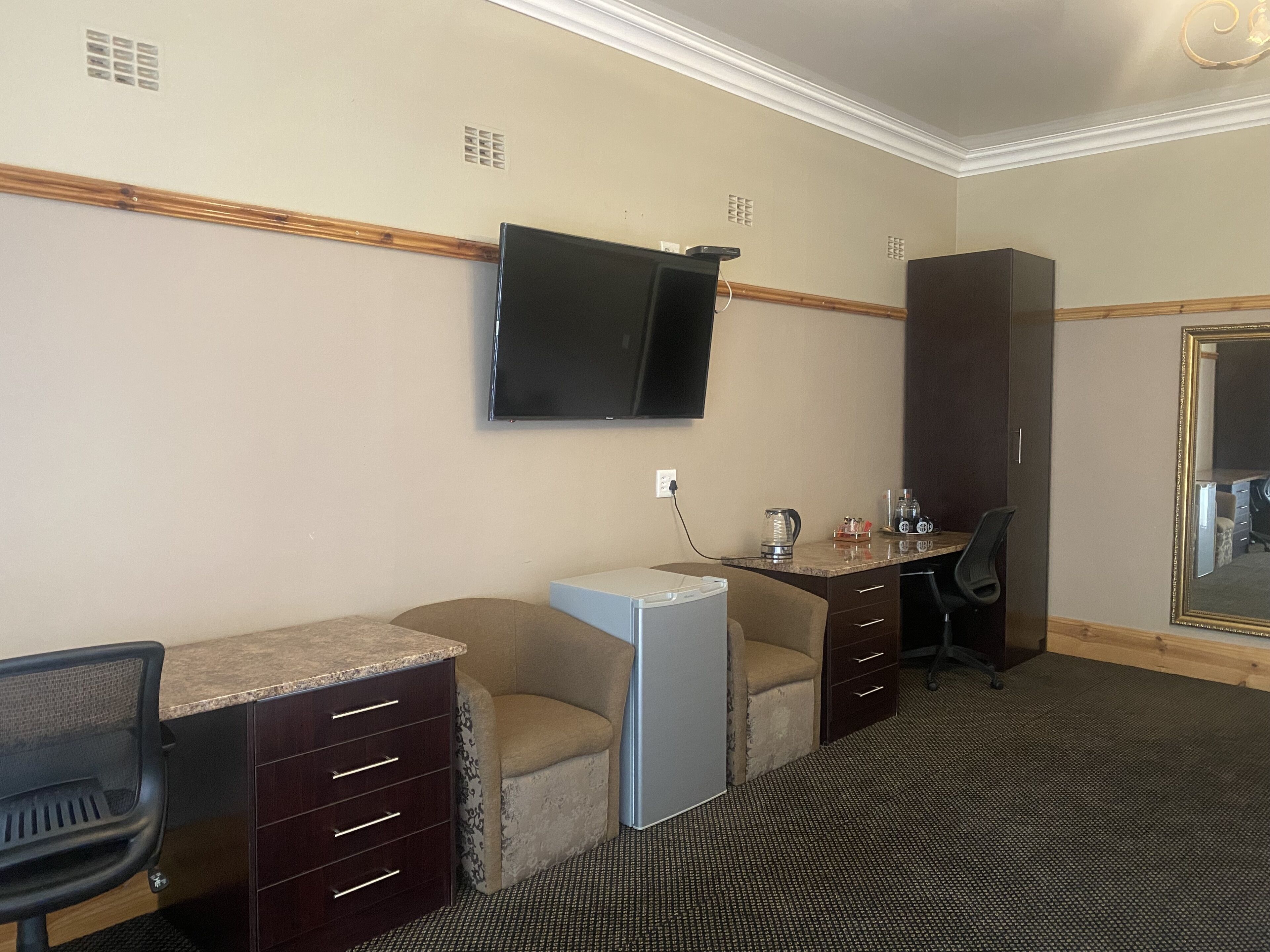 A hotel room with a desk, chair, kitchenette, and a television mounted on the wall.
