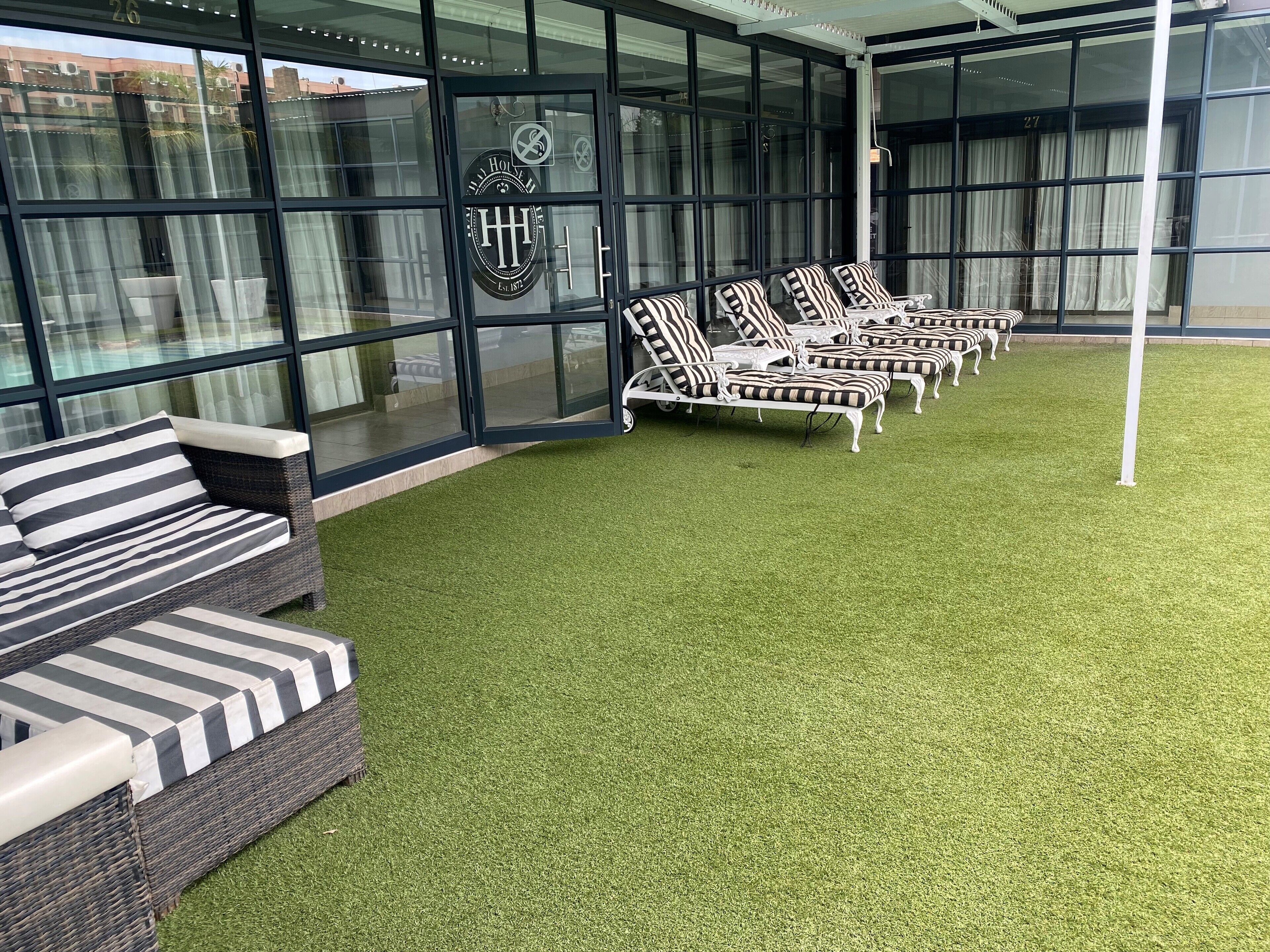 A hotel lobby with a glass facade, a logo on the door, and a row of striped lounge chairs on artificial grass.