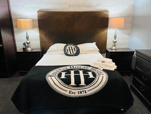 A neatly made bed with a black and white blanket featuring a logo and the establishment year. There are two bedside tables with lamps, a dark wooden headboard, and a black dresser. - The Halfway House Hotel (Kimberley)