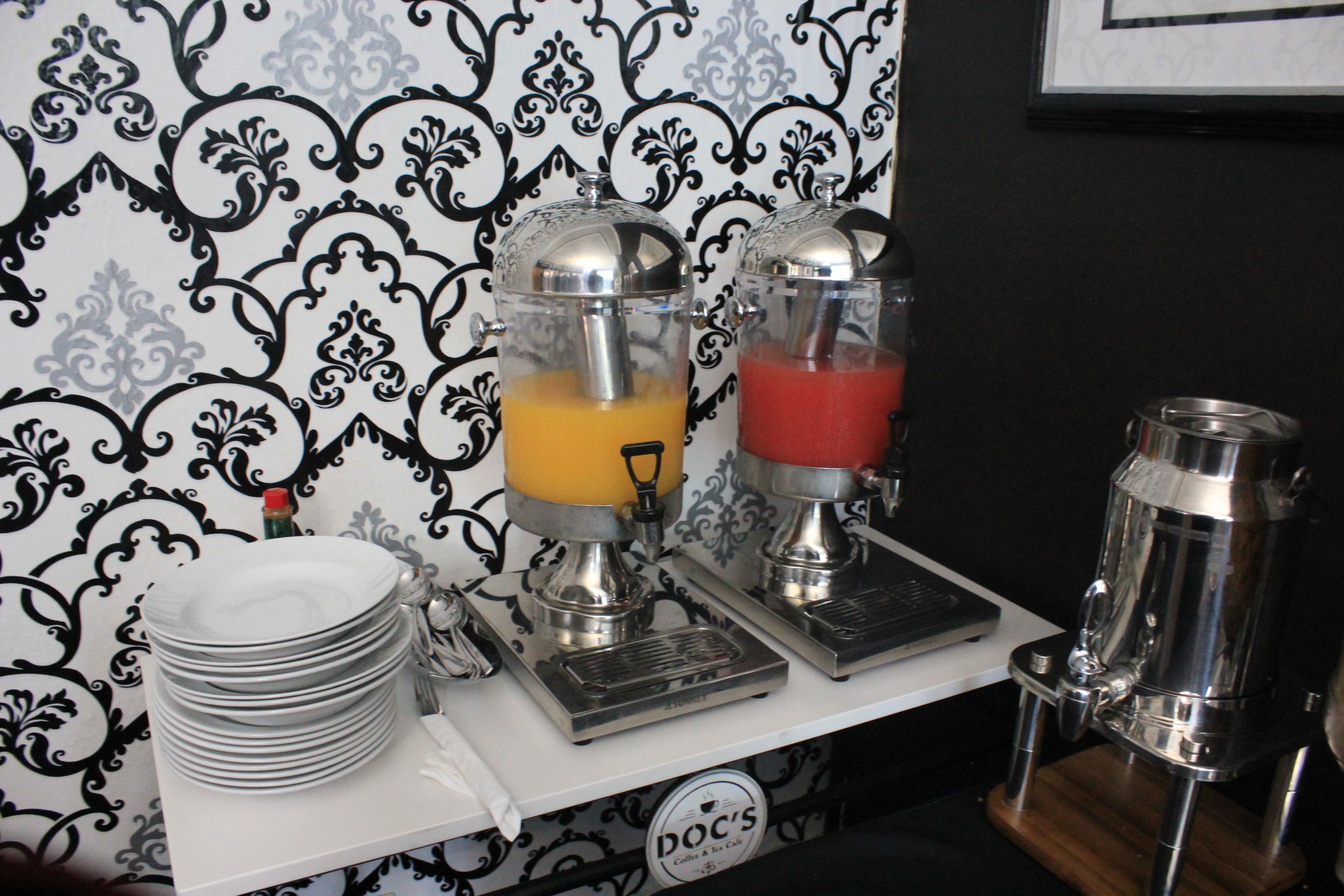 A beverage dispenser with a glass lid and a metal base, a red liquid container, and a stack of white plates on a white counter.