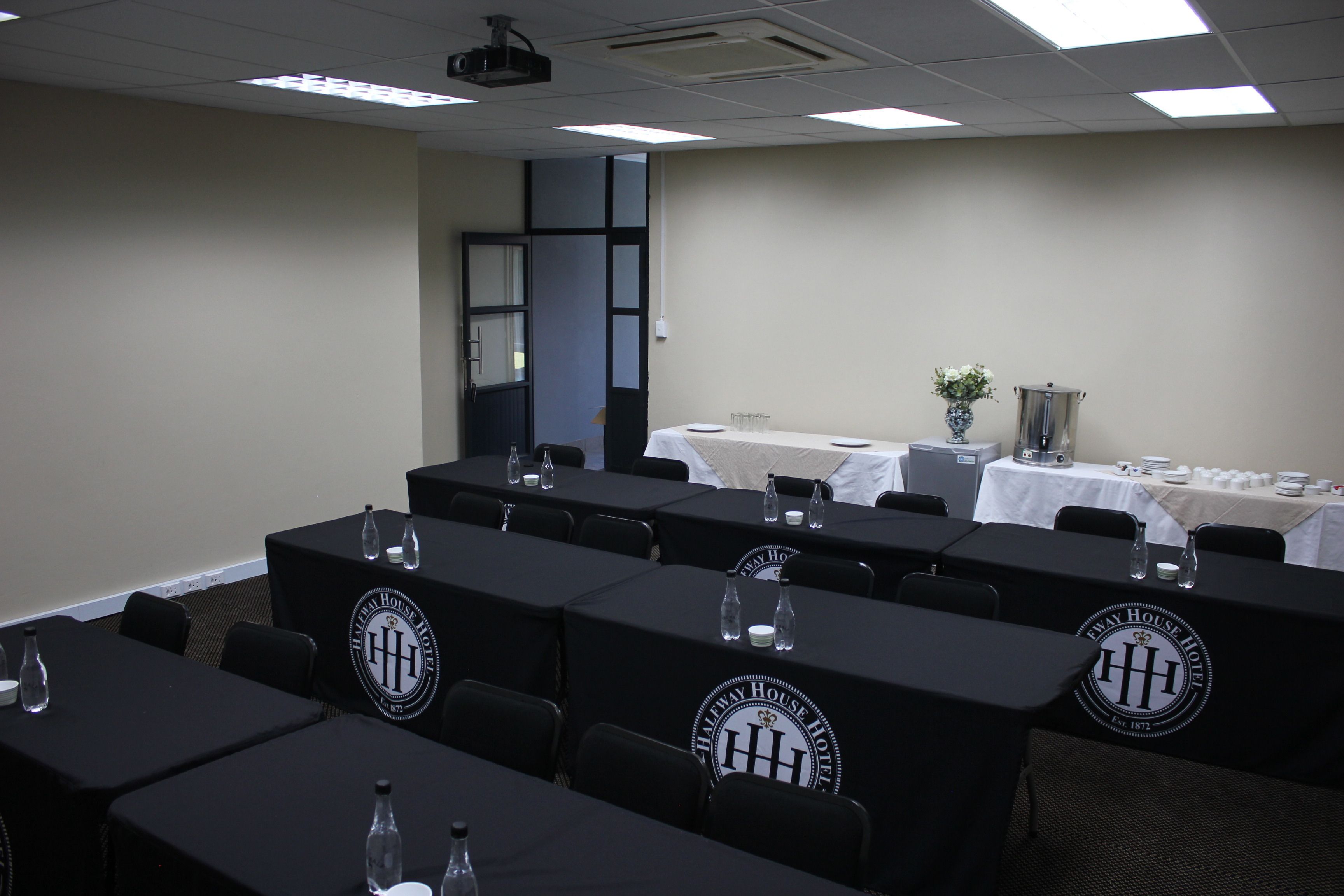 Un salón de conferencias con varias mesas, cada una cubierta con un mantel negro y un logotipo. Hay sillas alrededor de las mesas, y se ve una mesa blanca con un florero en el fondo.