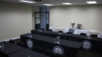 Un salón de conferencias con varias mesas, cada una cubierta con un mantel negro y un logotipo. Hay sillas alrededor de las mesas, y se ve una mesa blanca con un florero en el fondo.