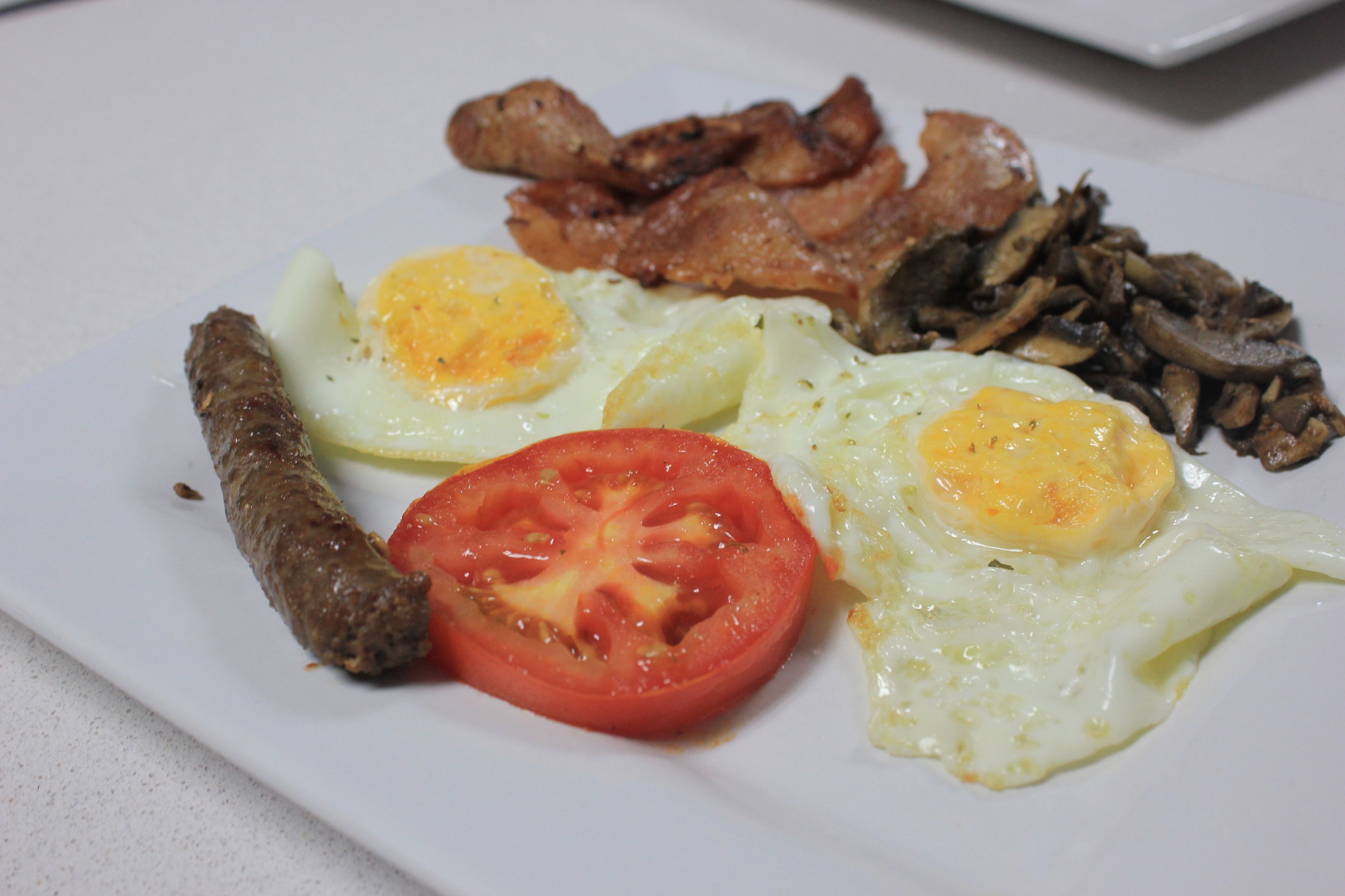 A plate of breakfast food including eggs, bacon, sausage, and a slice of tomato.