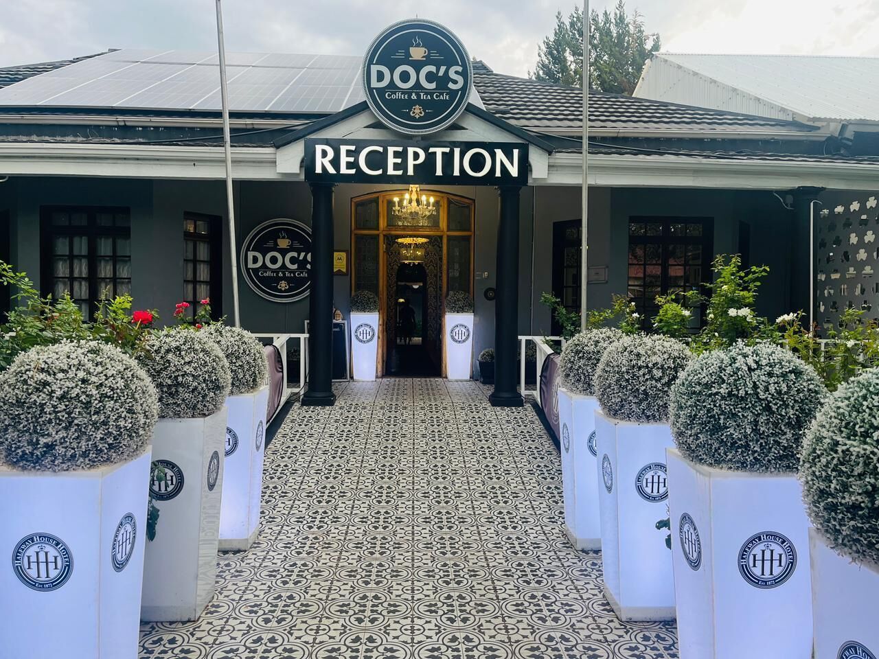 Un área de recepción con un letrero que dice 'Doc's Coffee & Tea Café', piso de baldosas y plantas en macetas.
