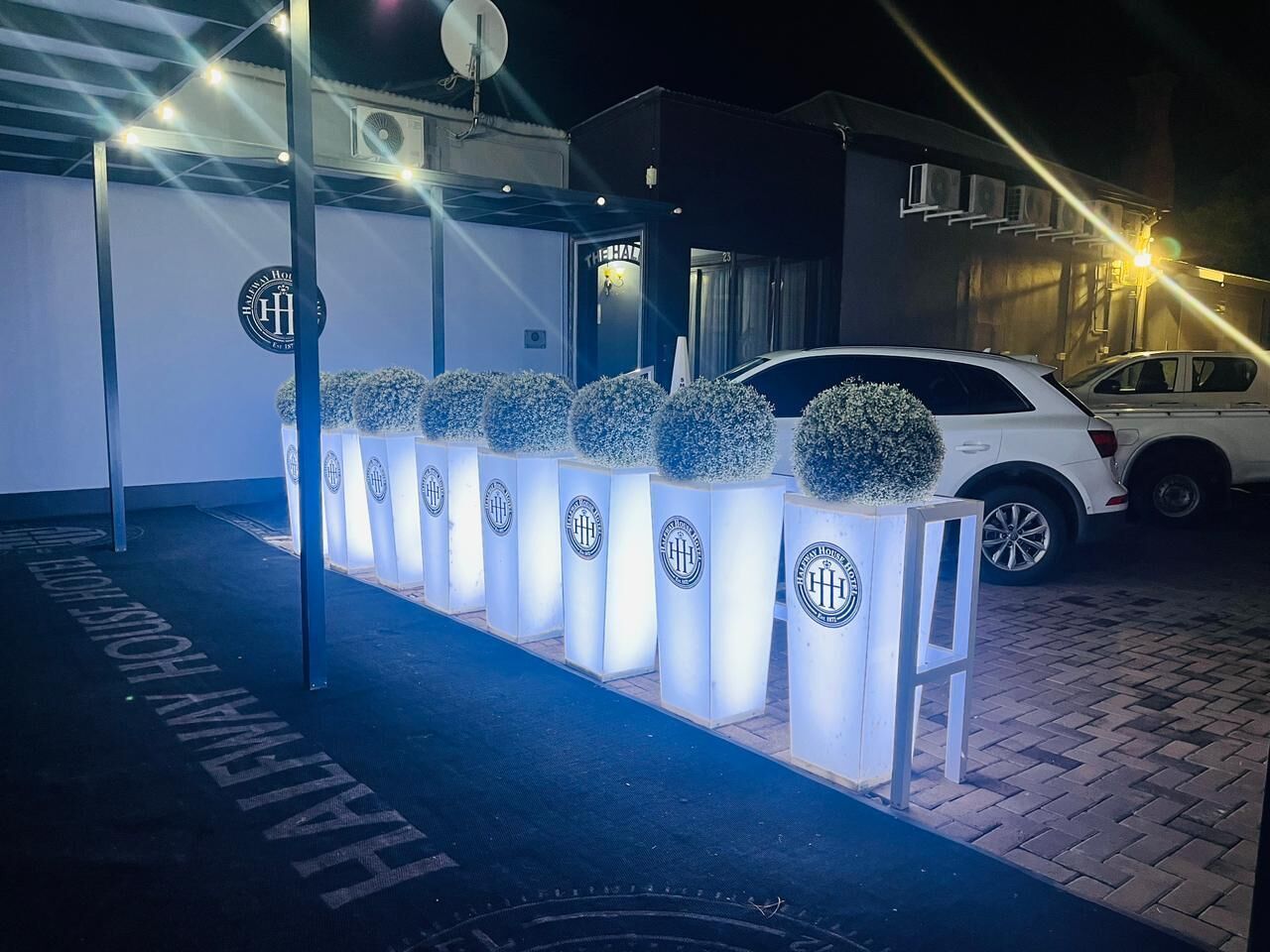 Une rangée de jardinières illuminées ornées d’un logo, une voiture blanche garée devant, et un bâtiment avec une enseigne en arrière-plan.