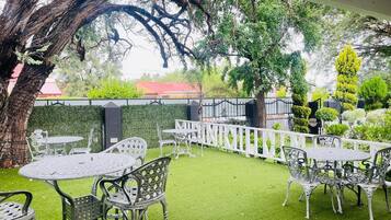 An outdoor patio area with tables and chairs, surrounded by a white fence and greenery.