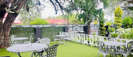 Un espace extérieur aménagé en patio, avec des tables et des chaises, entouré d’une clôture blanche et de verdure.