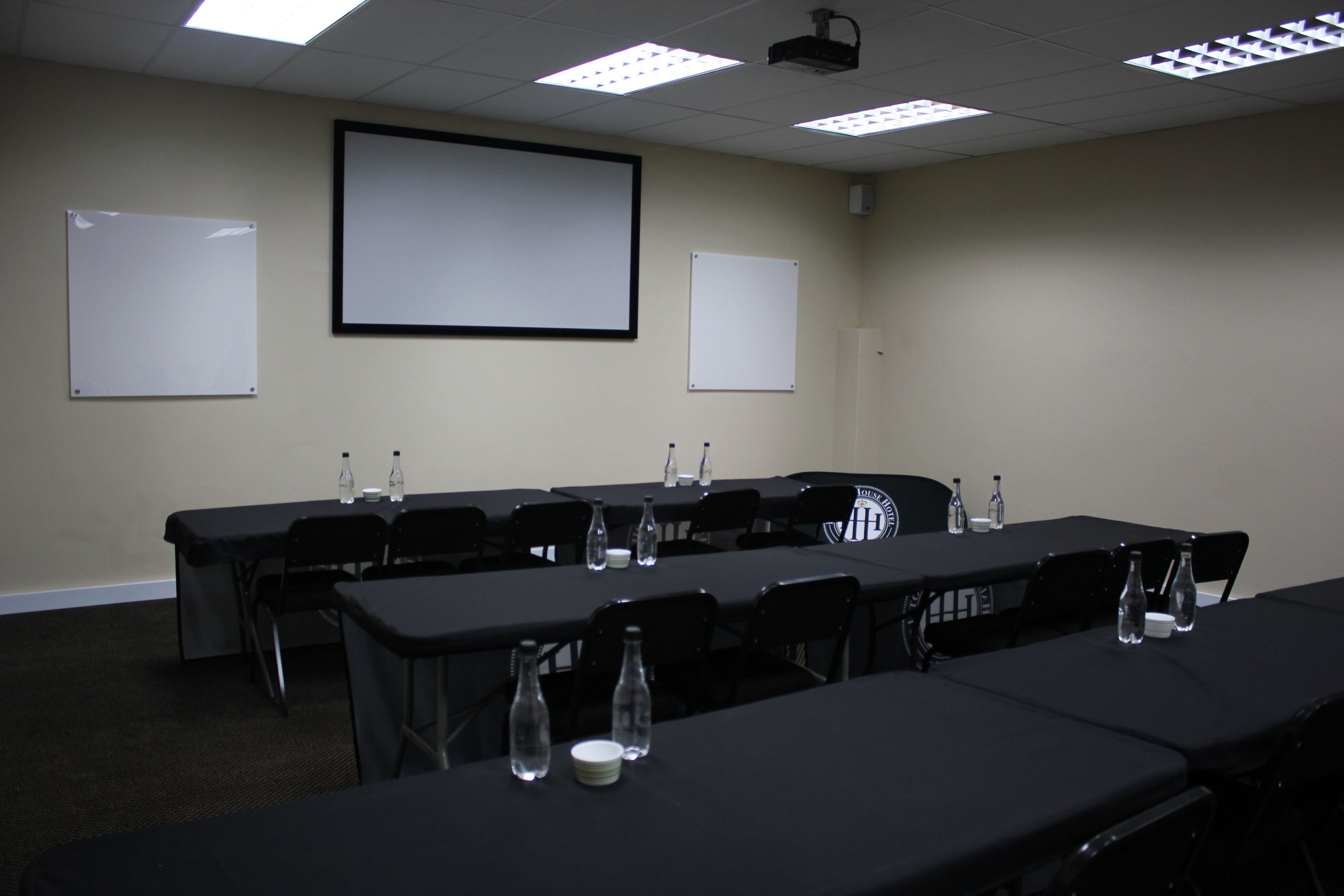 A conference room with a projector screen, whiteboards, tables, chairs, and water bottles.