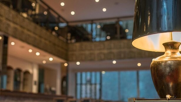 A modern interior with a large, ornate lamp on a table, a high ceiling, and a mezzanine level with seating. - America Hotel (Mar del Plata)