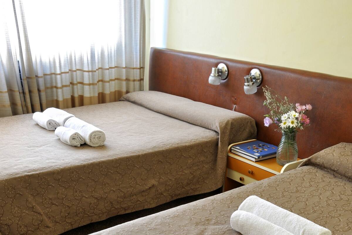 Two twin beds with beige patterned bedspreads, a wooden headboard, and a small bedside table with a book and a vase of flowers.