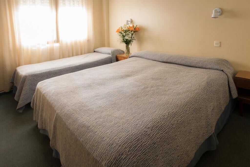 A hotel room with two single beds, a nightstand, a vase with flowers, and a wall-mounted lamp.