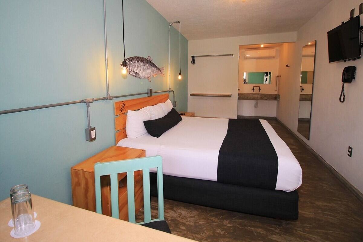 Habitación de hotel con cama, mesita de noche de madera, una silla, una pequeña mesa, decoración de pez, espejo, teléfono y un baño visible al fondo.