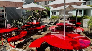 An outdoor seating area with red tables and beige umbrellas, surrounded by greenery and a stone wall.