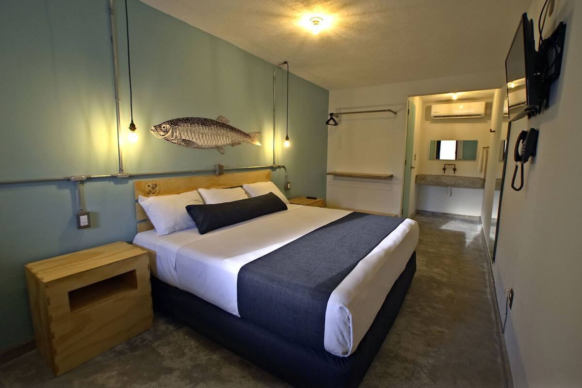 Un dormitorio con cama, mesitas de noche, un adorno de pez en la pared, una lámpara colgante y un baño visible a través de una puerta abierta.