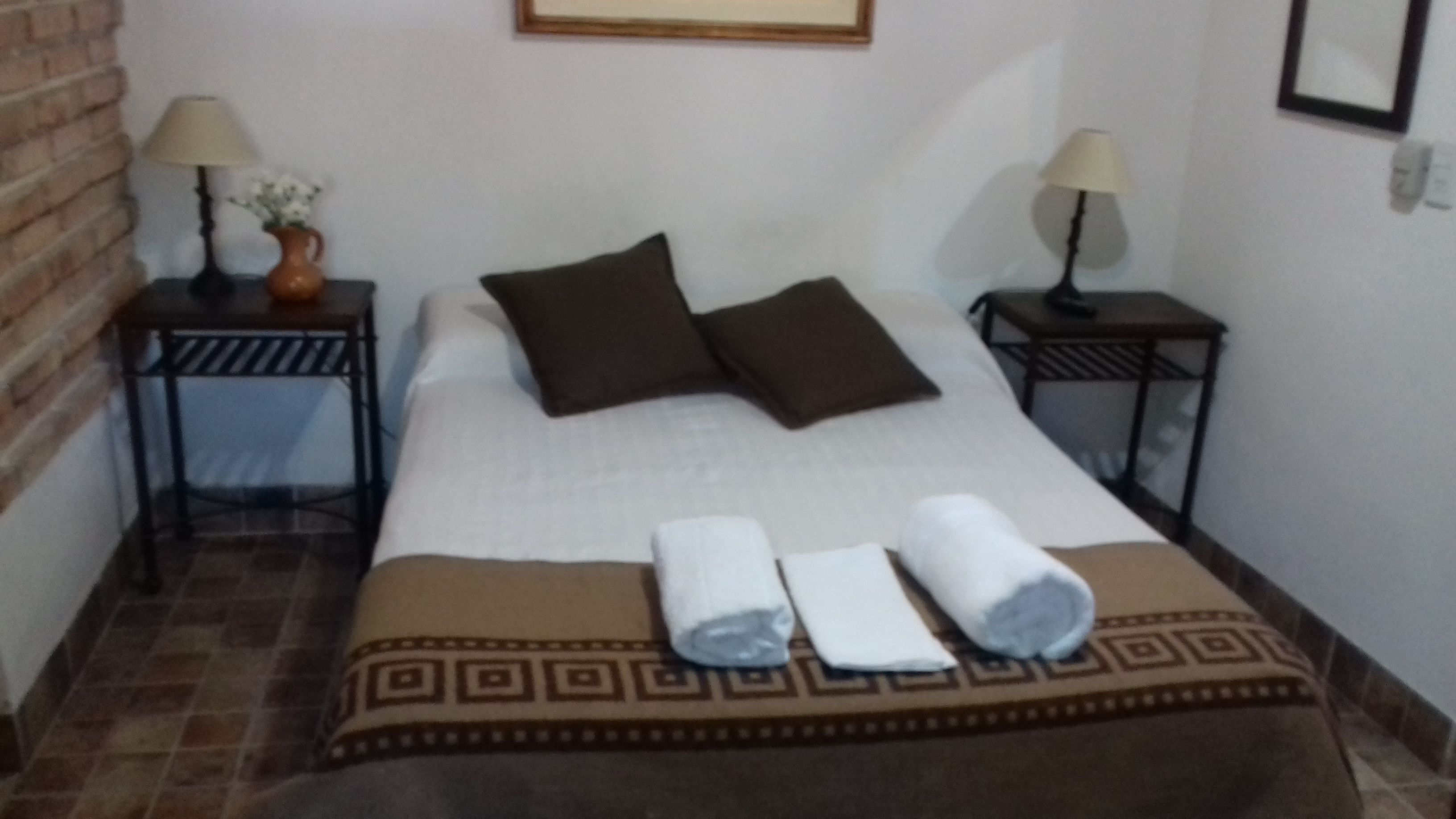 A neatly made bed with a patterned bedspread, two pillows, and two rolled towels. There are two bedside tables with lamps and a vase of flowers on one.