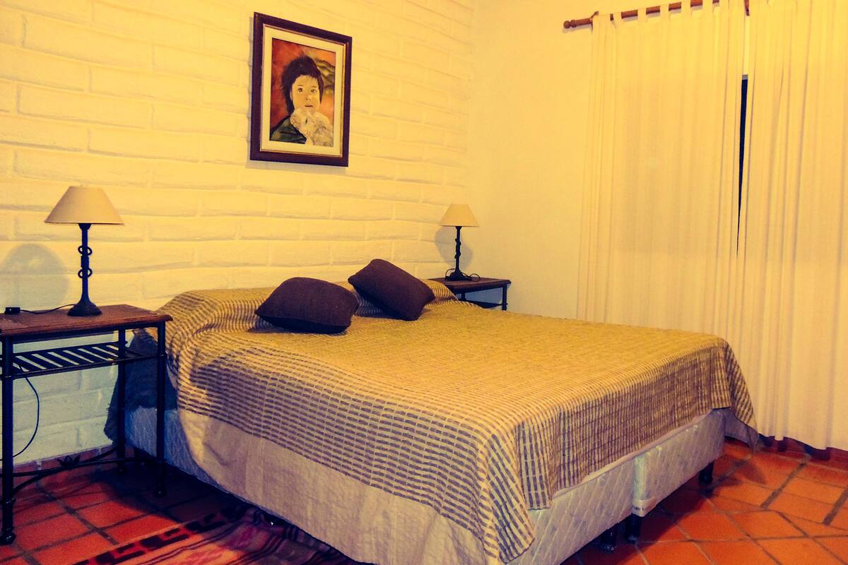 A neatly made bed with a striped bedspread and two pillows, flanked by nightstands with lamps, in a room with a white brick wall and a framed picture above the bed.