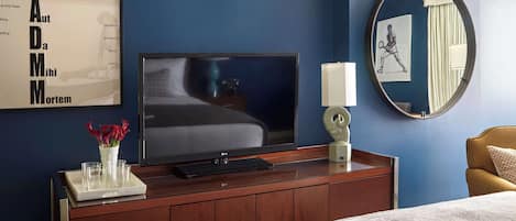 Una habitación de hotel con una televisión sobre un mueble de madera, una mesita de noche con lámpara, un espejo y un cuadro en la pared.