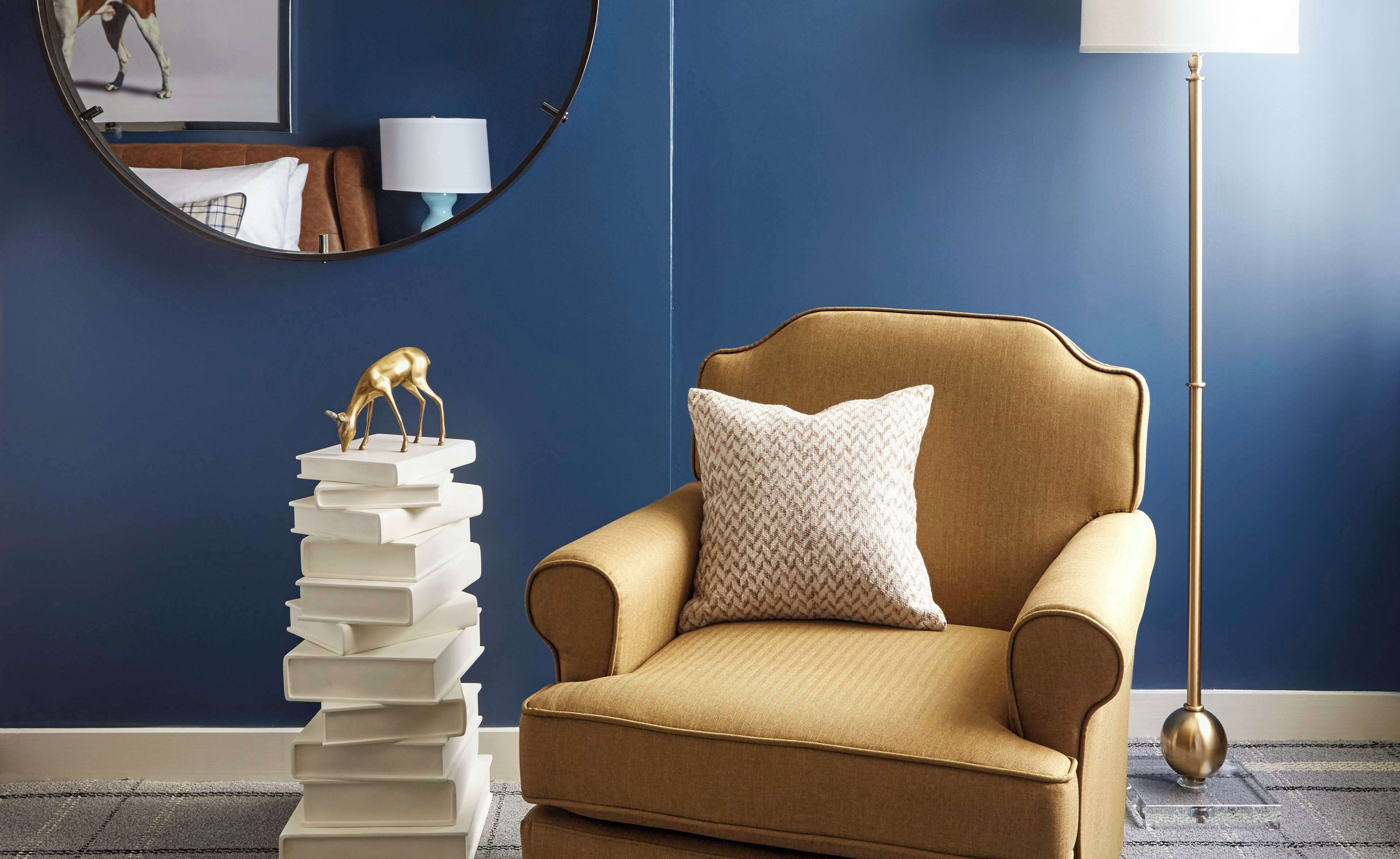 A room with a blue wall, a gold armchair with a patterned pillow, a stack of books with a golden deer figurine on top, a round mirror, and a floor lamp.