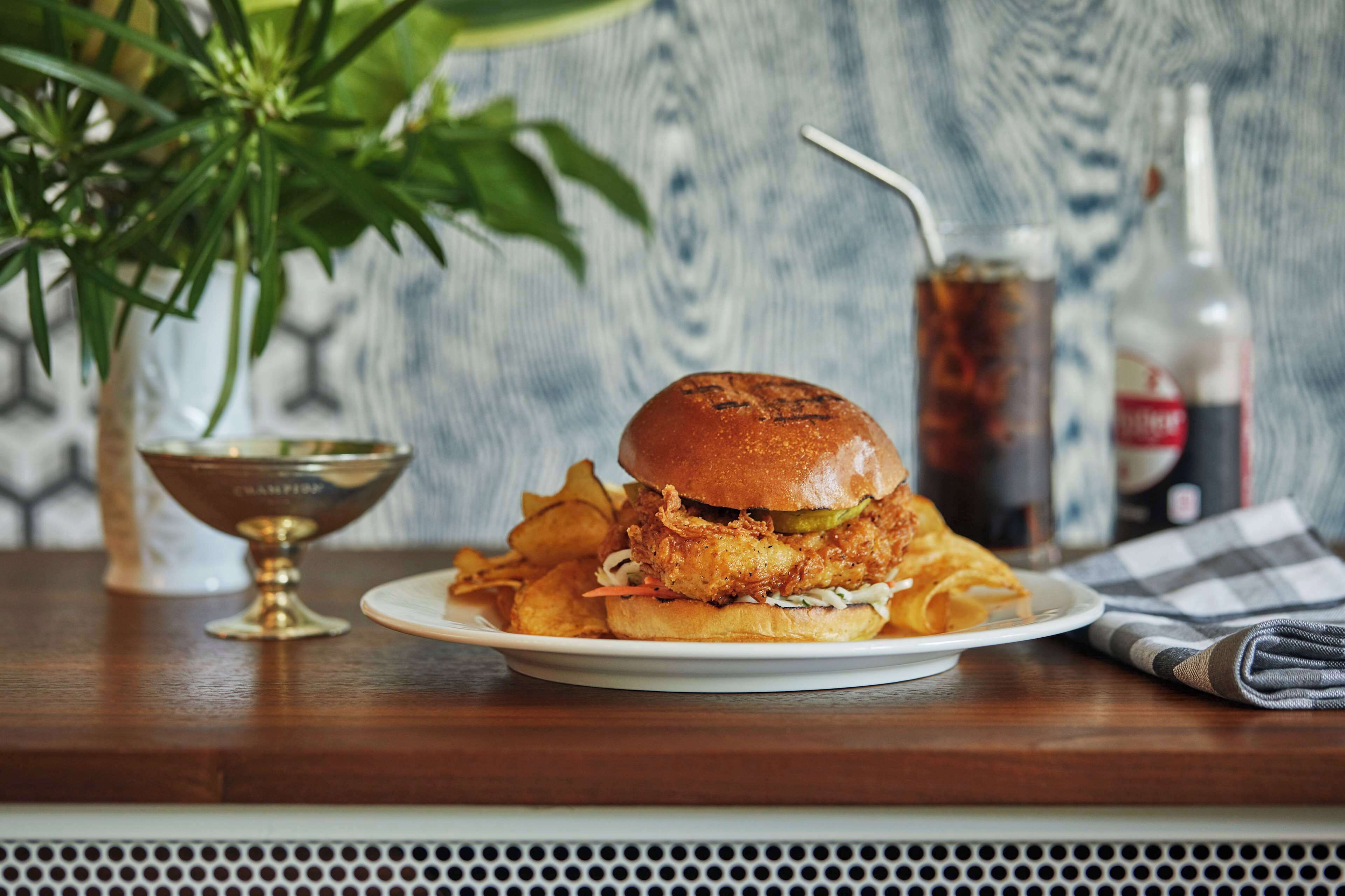 Un plato con un sándwich de pollo frito y papas fritas, un vaso de gaseosa y una botella de salsa sobre una mesa de madera.