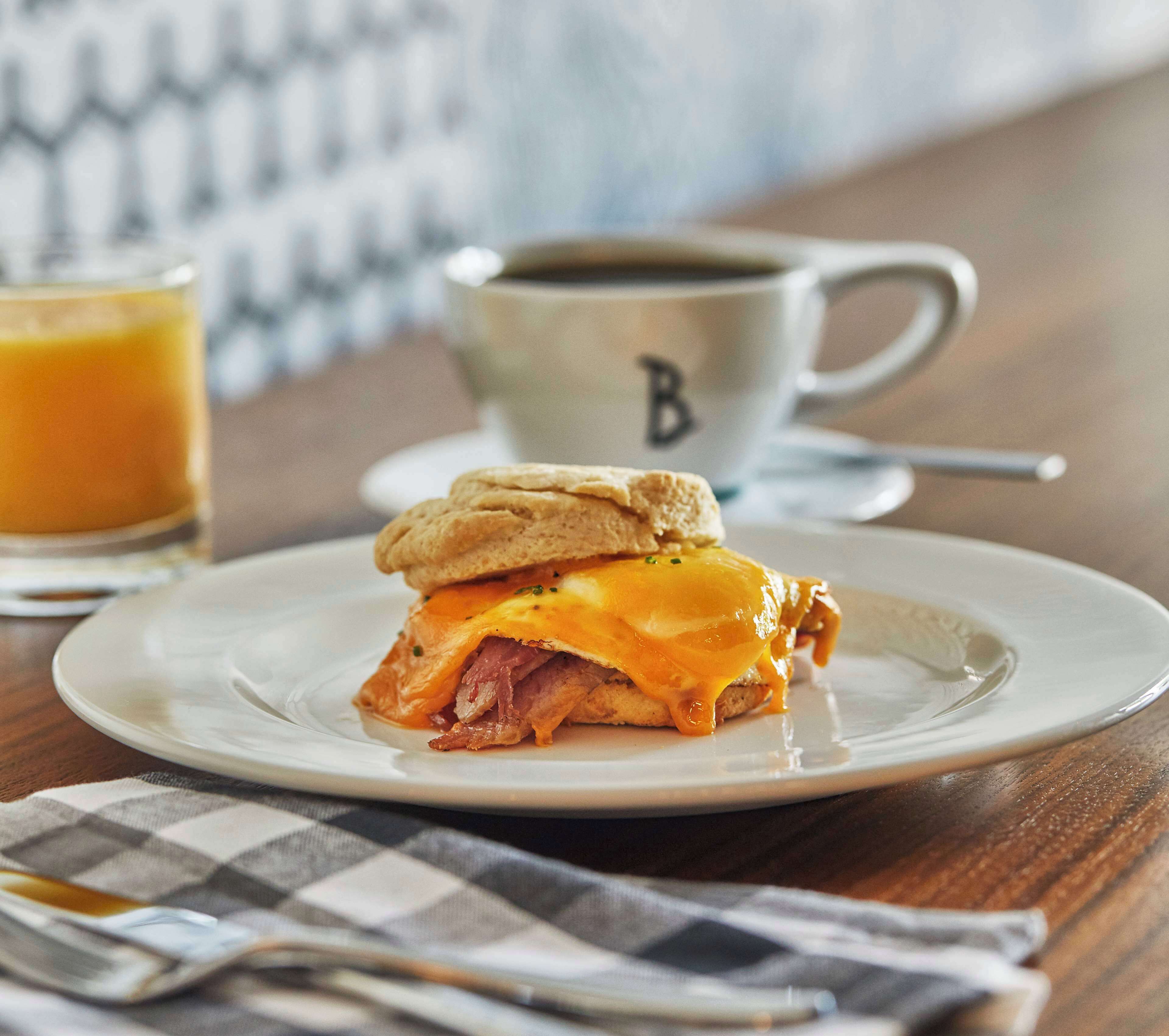 A breakfast sandwich on a plate with a cup of coffee and a glass of orange juice in the background.