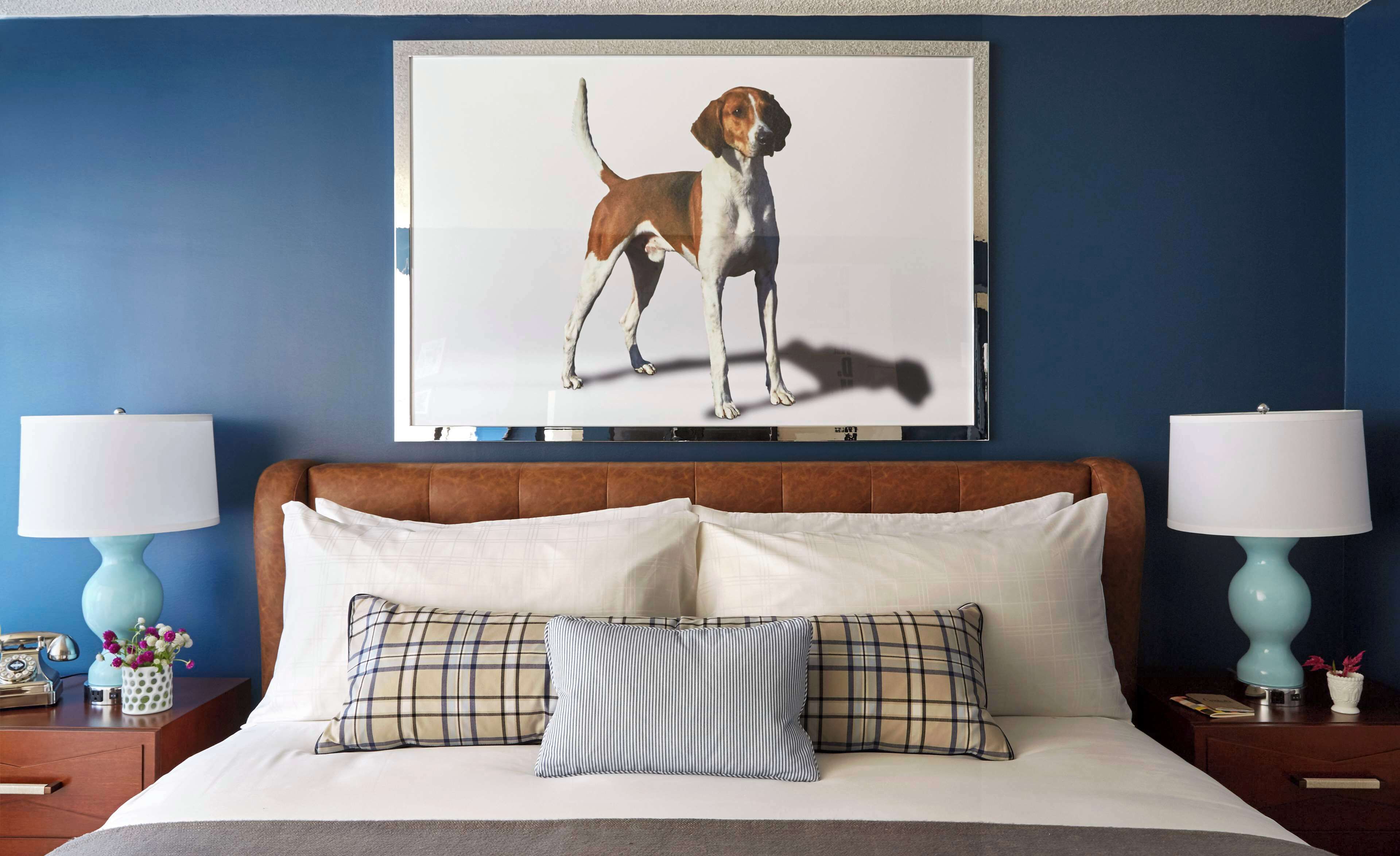 A bedroom with a large bed, two bedside tables with lamps, a framed picture of a dog on the wall, and a blue wall.