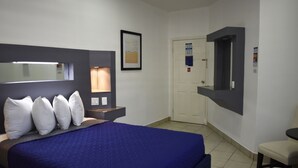 A hotel room with a large bed, a bedside table, a chair, a desk, a mirror, and a door. - Hotel Mediterraneo (Playas de Rosarito)