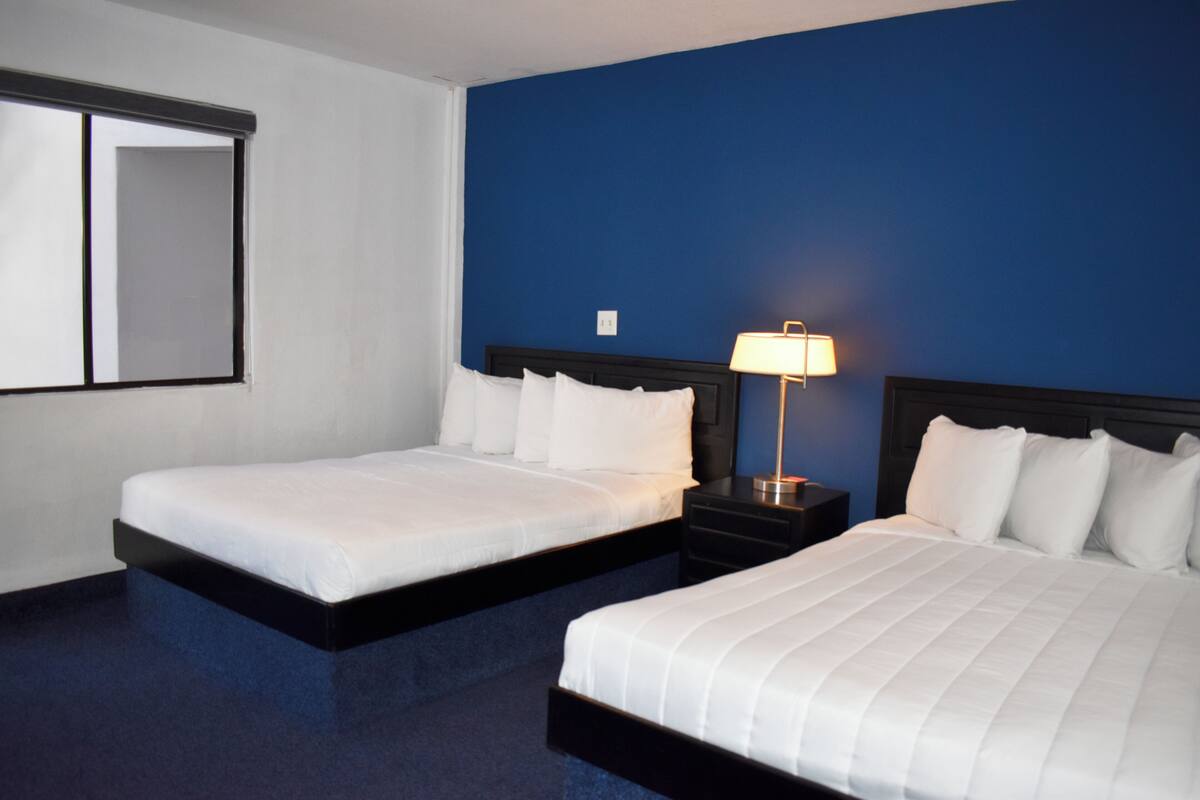 A hotel room with two beds, a nightstand with a lamp, and a large window.
