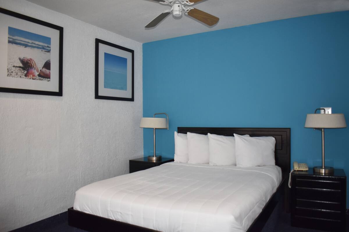 A hotel room with a bed, two nightstands, two lamps, a ceiling fan, and two framed pictures on the wall.