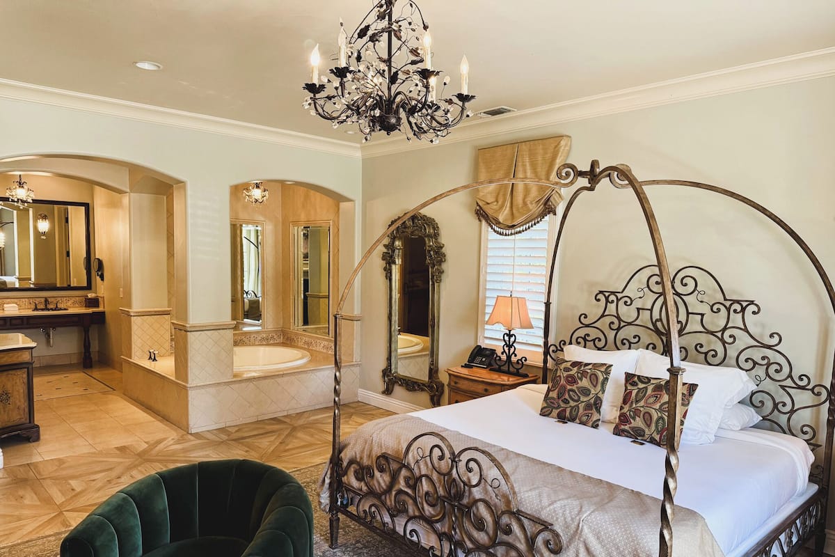 Ein geräumiges Schlafzimmer mit Himmelbett, einer Sitzecke inklusive grünem Ohrensessel, einem großen Spiegel, einer Chandelierleuchtre und einem Badezimmer mit Badewanne und Waschtisch.