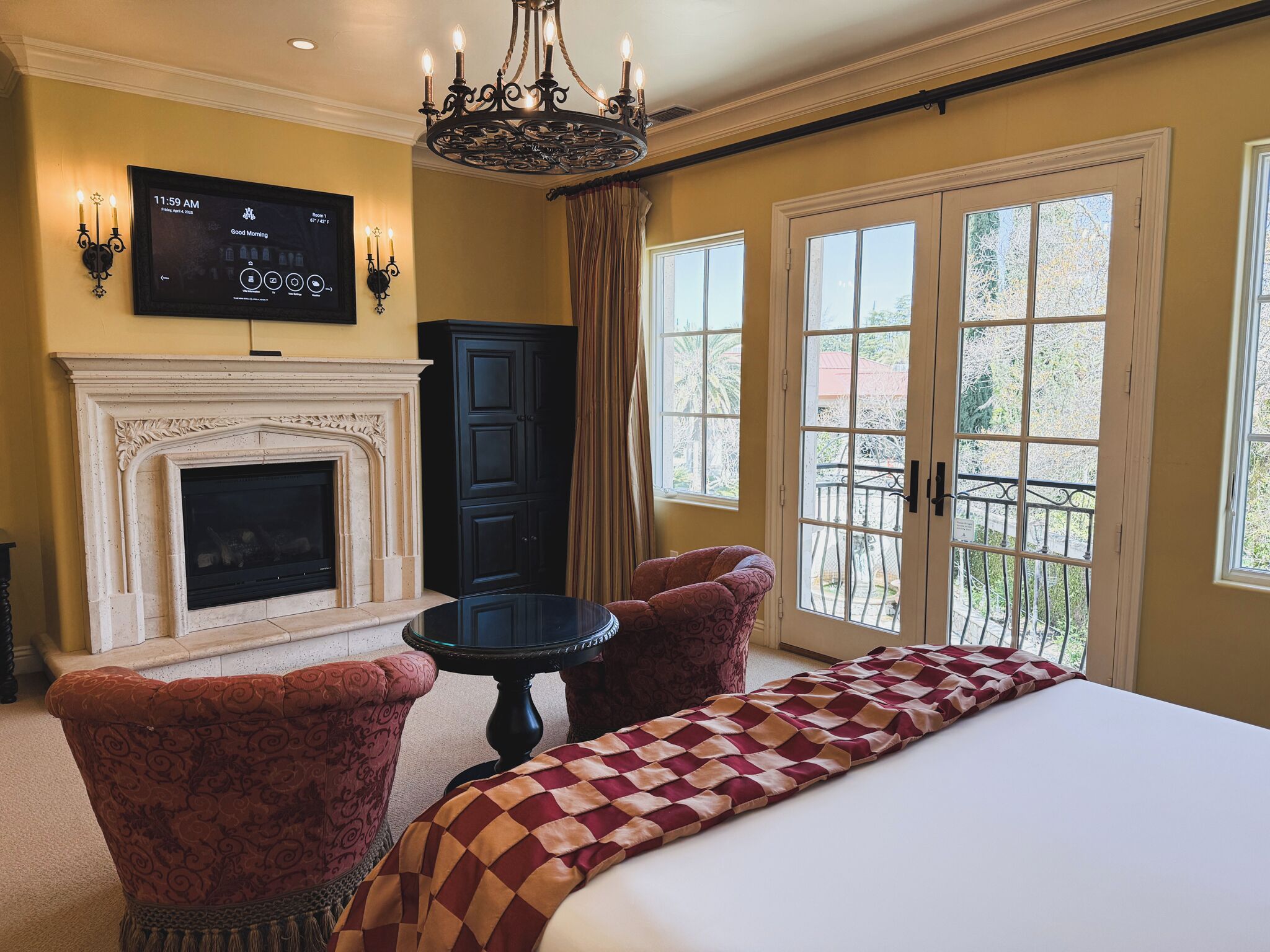 A hotel room with a large bed, two armchairs, a fireplace, a television, and a balcony with a view.