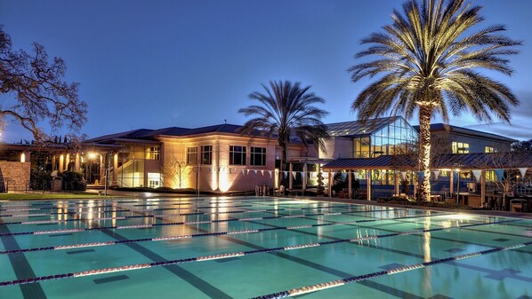 A large outdoor swimming pool with multiple lanes, surrounded by a well-lit building with a covered patio area and palm trees. - Villa Arden Hills (Sacramento)