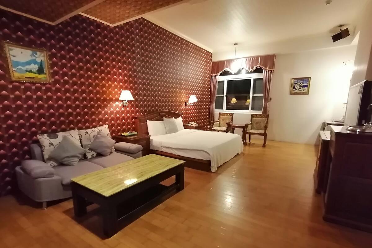 A hotel room with a bed, sofa, and armchair. There is a wooden coffee table and a desk with a chair. The room has a patterned wall and a window with curtains.