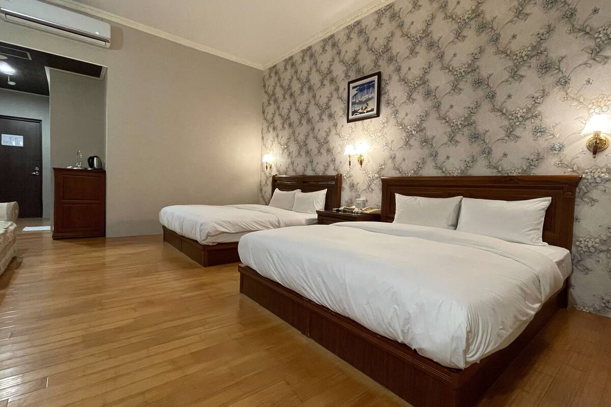 A hotel room with two beds, wooden headboards, a wooden dresser, a desk with a lamp, and a floral wallpaper.