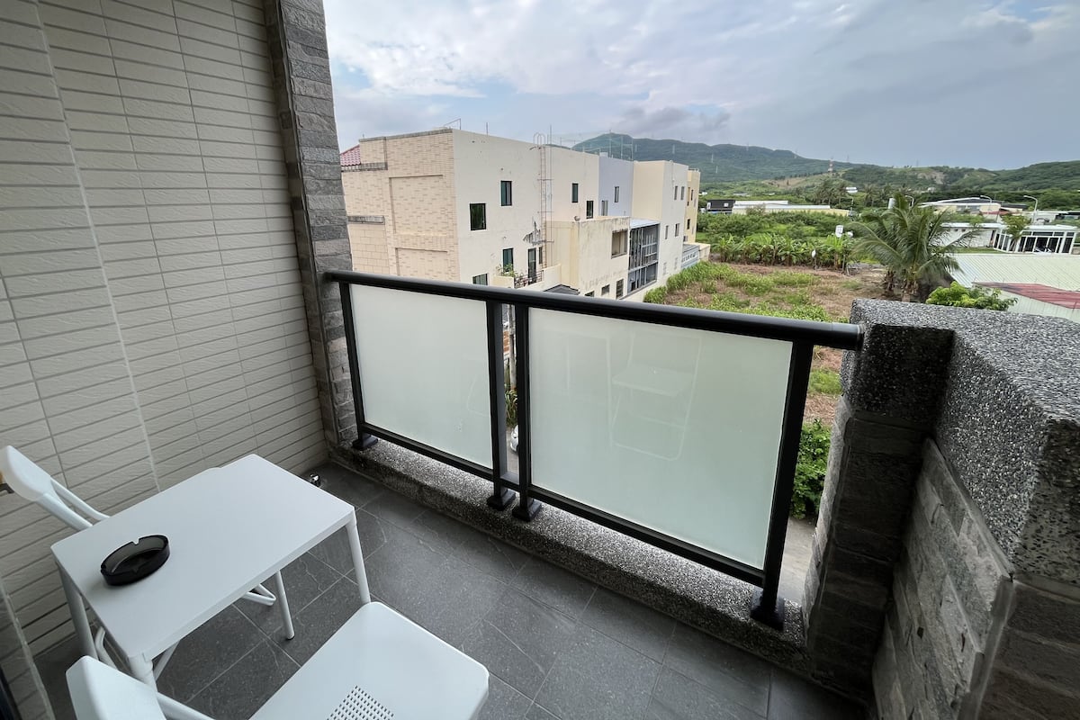 A balcony with a white table and chair, a glass railing, and a view of buildings and greenery.