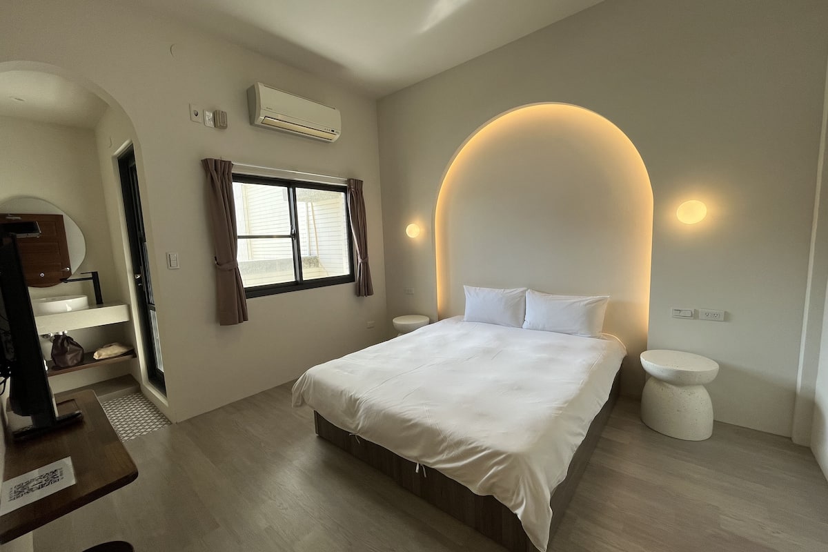 A modern bedroom with a large bed, a window with curtains, a built-in air conditioner, and a unique curved wall light.
