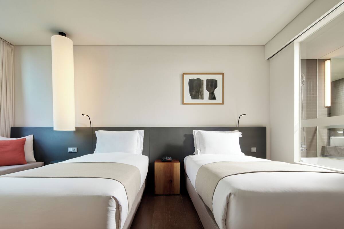 A hotel room with two beds, a nightstand, a framed artwork on the wall, and a window with a view of the bathroom.