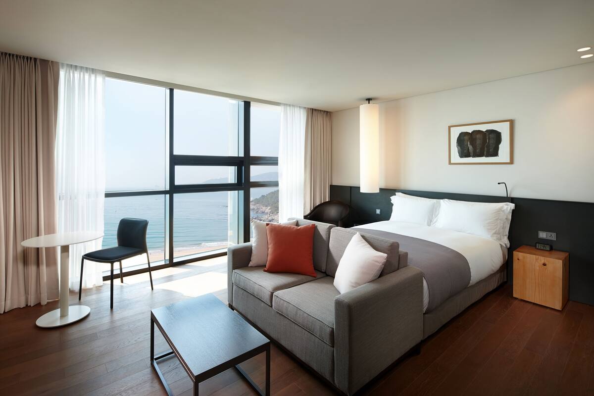 A modern hotel room with a large bed, a sofa, a small table, and a view of the sea.