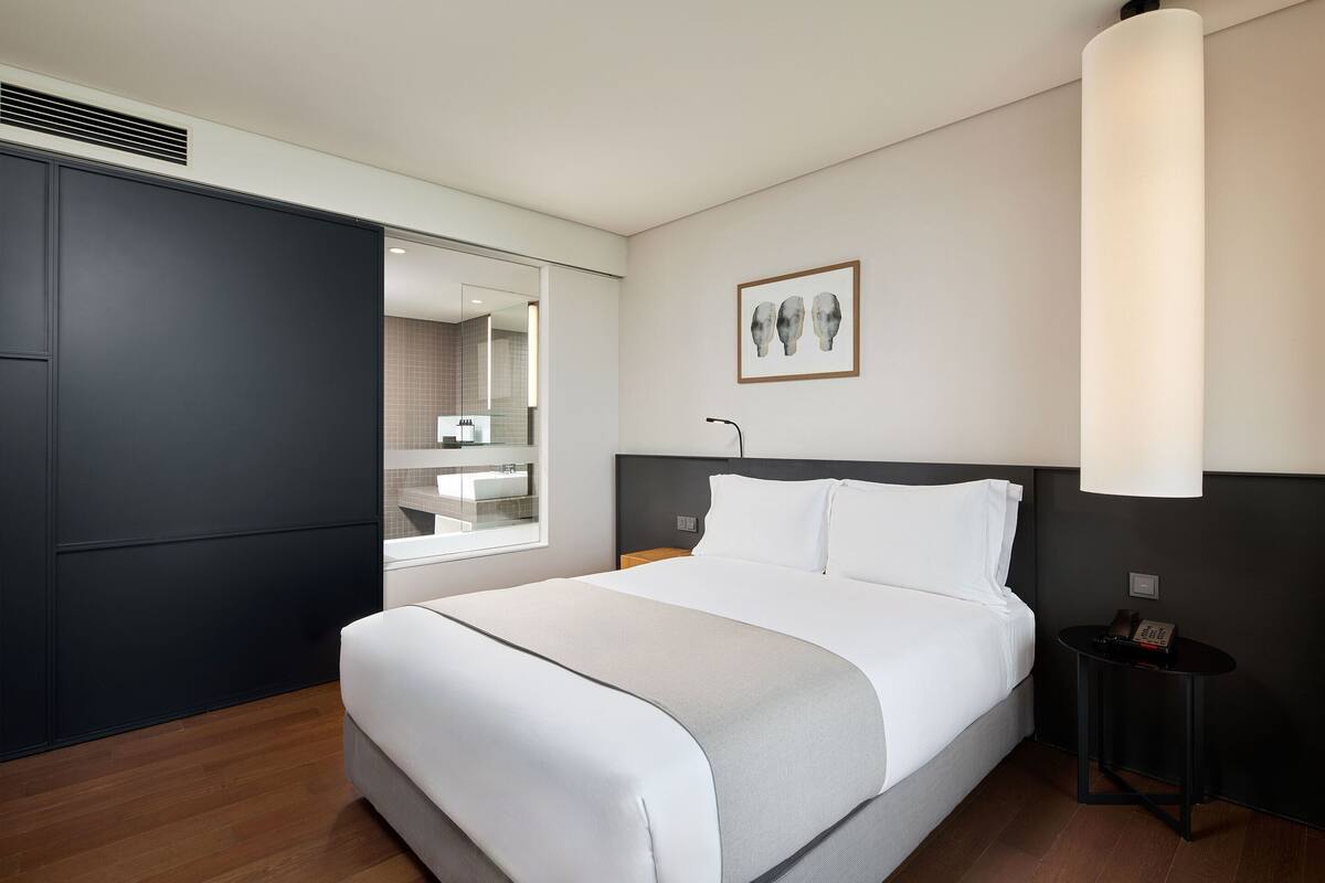 A modern hotel room with a large bed, a bedside table, a nightstand, and a large window.