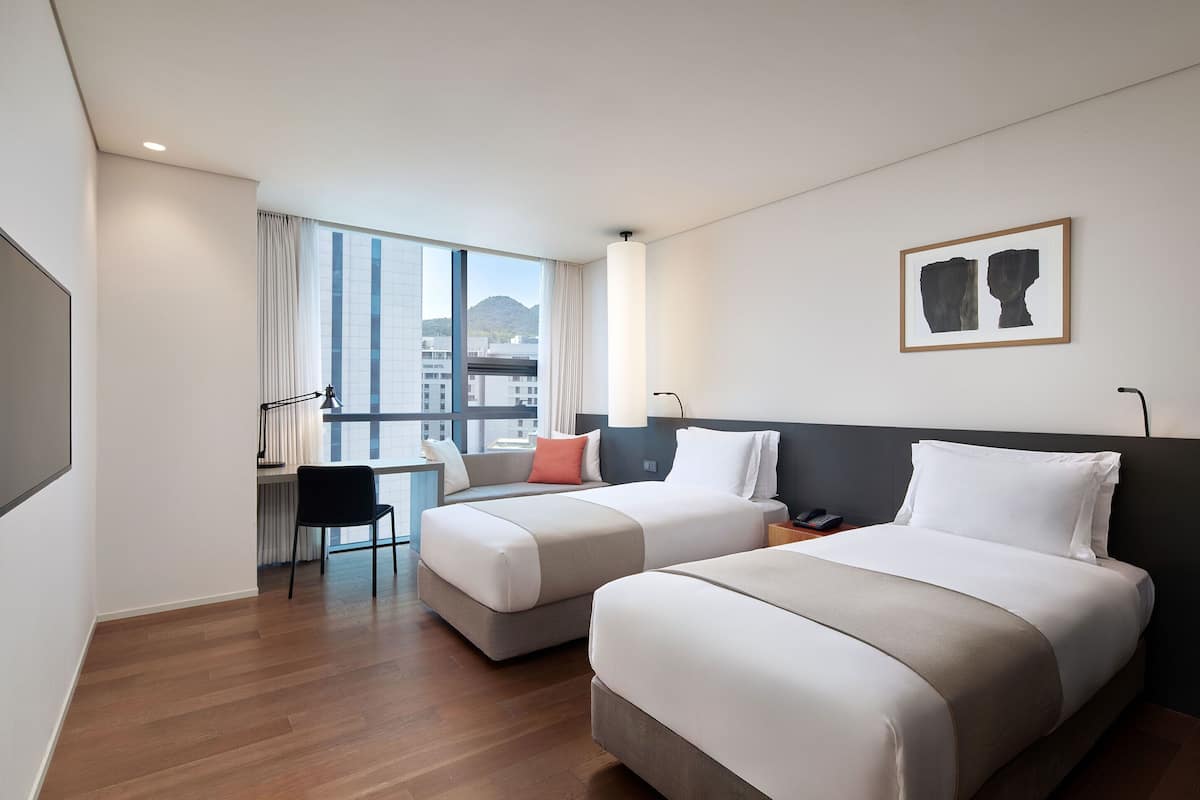 A hotel room with two beds, a desk, and a large window offering a city view.