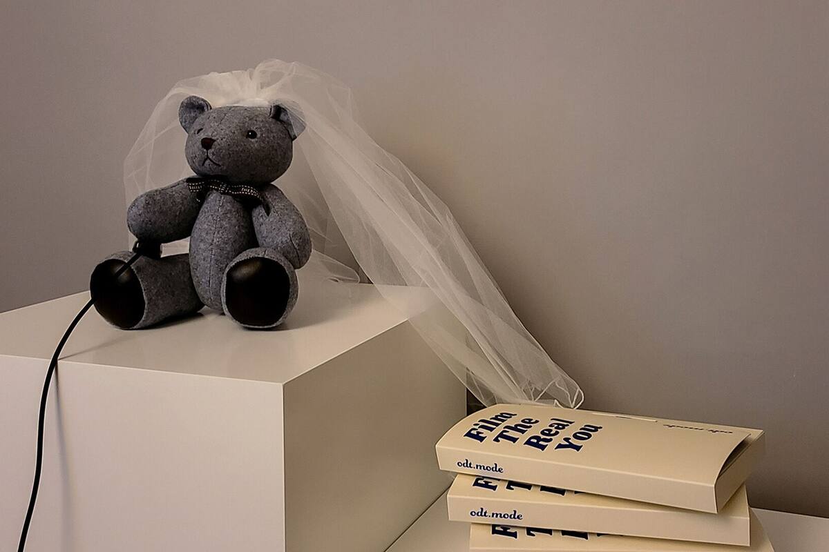 A grey teddy bear with a veil on a white surface, next to a stack of books with the text "Film The Real You".