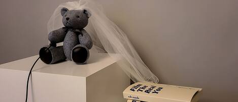A grey teddy bear with a veil on a white surface, next to a stack of books with the text "Film The Real You".
