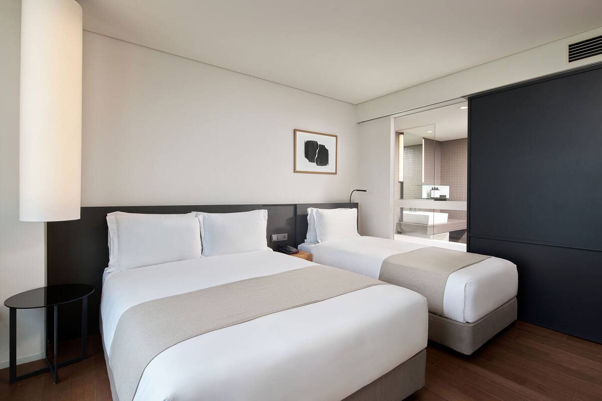 A hotel room with two beds, a bedside table, a lamp, a mirror, and a framed picture on the wall.